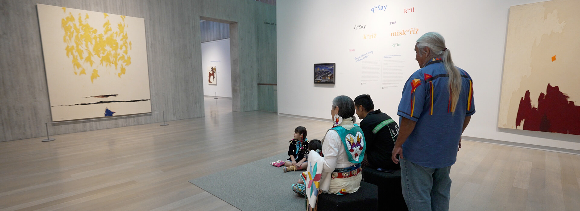 A family stands and sits while looking at an abstract painting with fluttery yellow shapes and dark horizontal lines