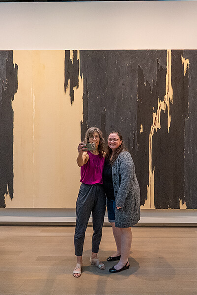 Two women take a selfie in front of a large painting