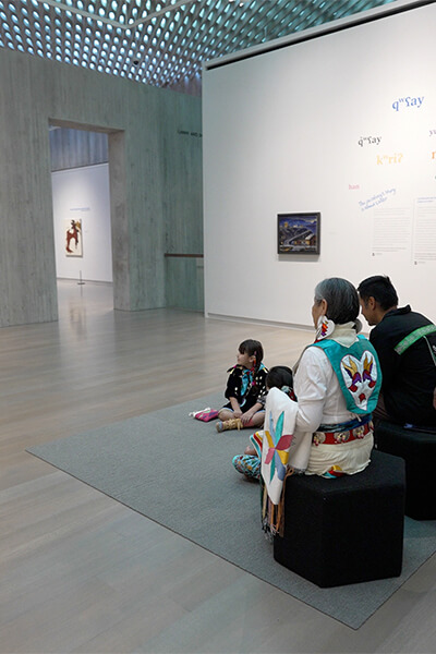 A family stands and sits while looking at an abstract painting with fluttery yellow shapes and dark horizontal lines