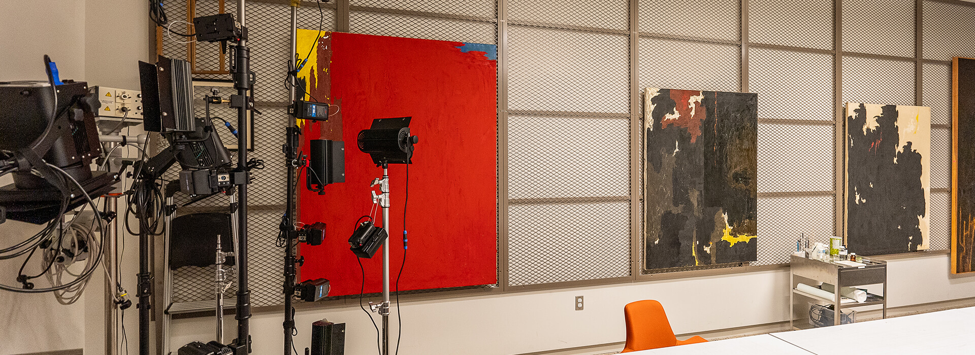 Paintings hang on racks with light stands in front of them in the conservation lab