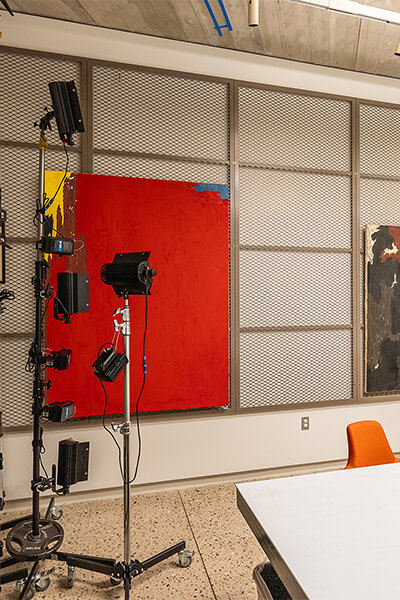 Paintings hang on racks with light stands in front of them in the conservation lab