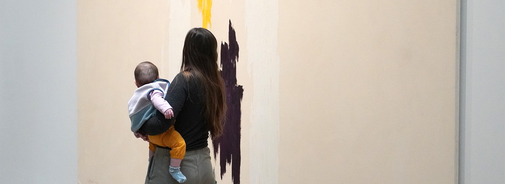 A woman holds her baby up on her hip as they look at a large abstract painting together