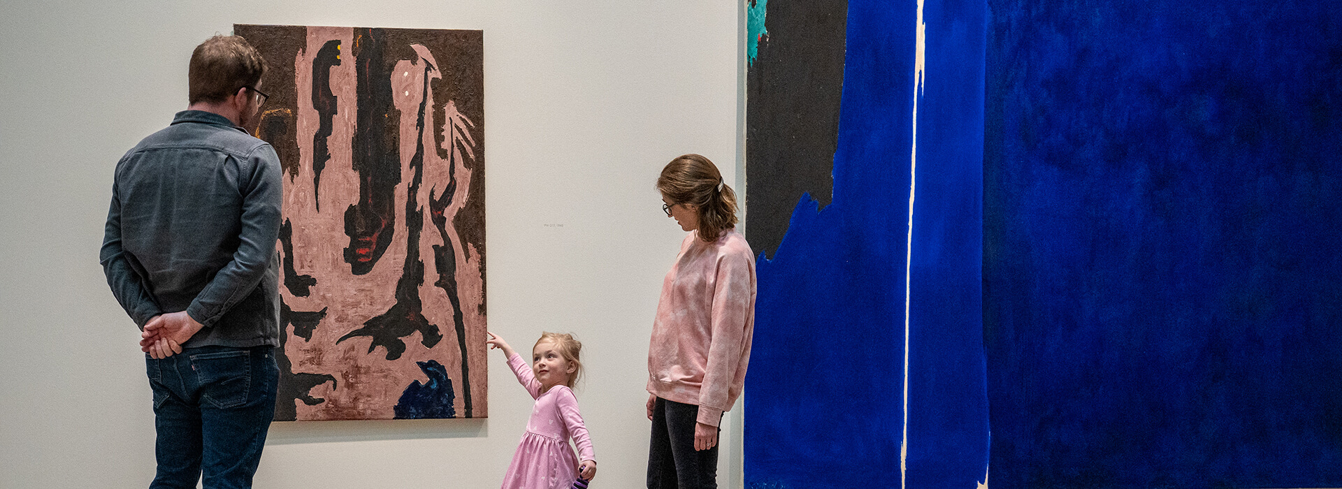 A young child points at a painting and looks up at her parents