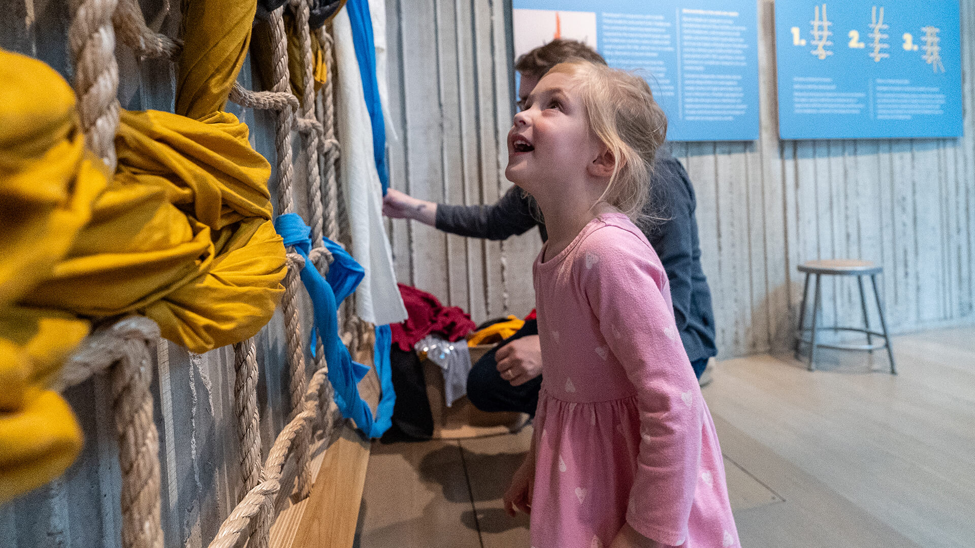 A young child looks up and smiles at a big weaving wall