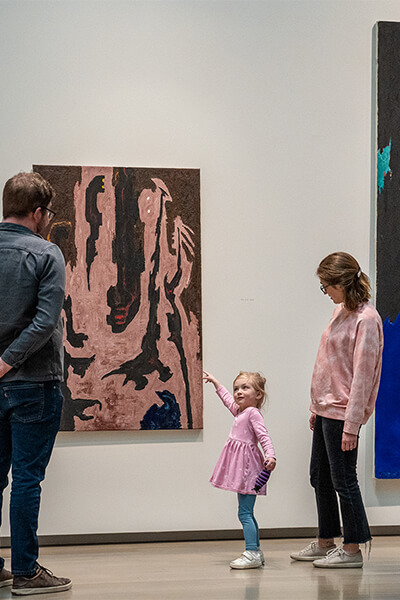 A young child points at a painting and looks up at her parents