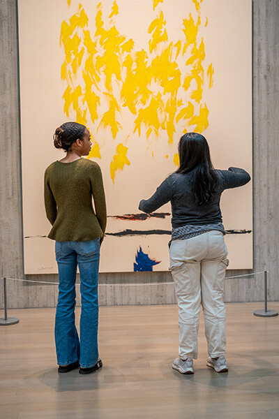 Two women look at a large yellow abstract painting