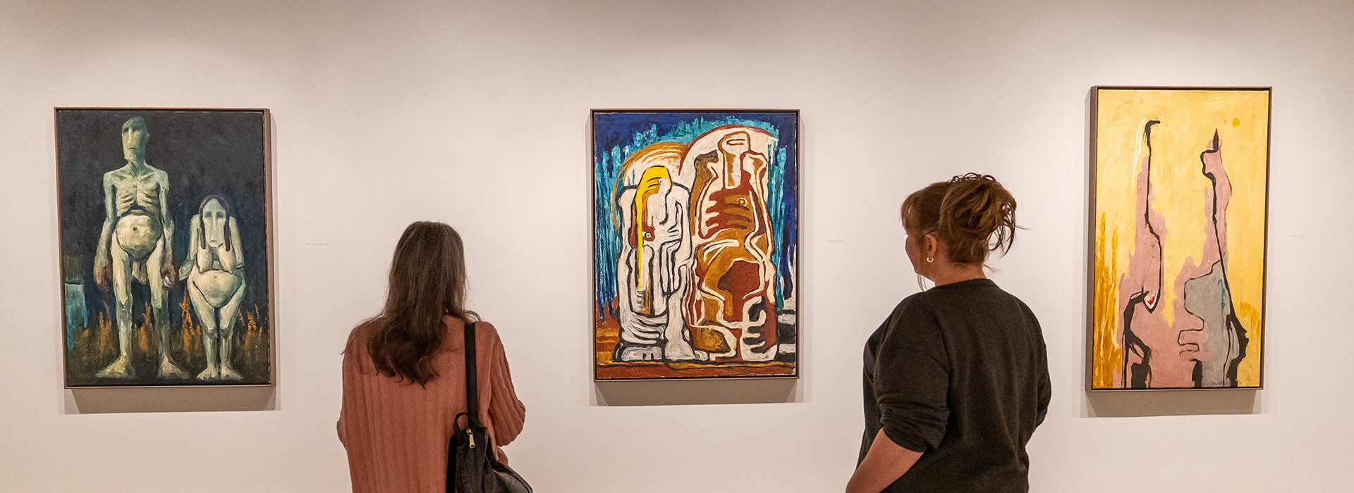 Two women look at an abstract painting of two people on a wall of paintings
