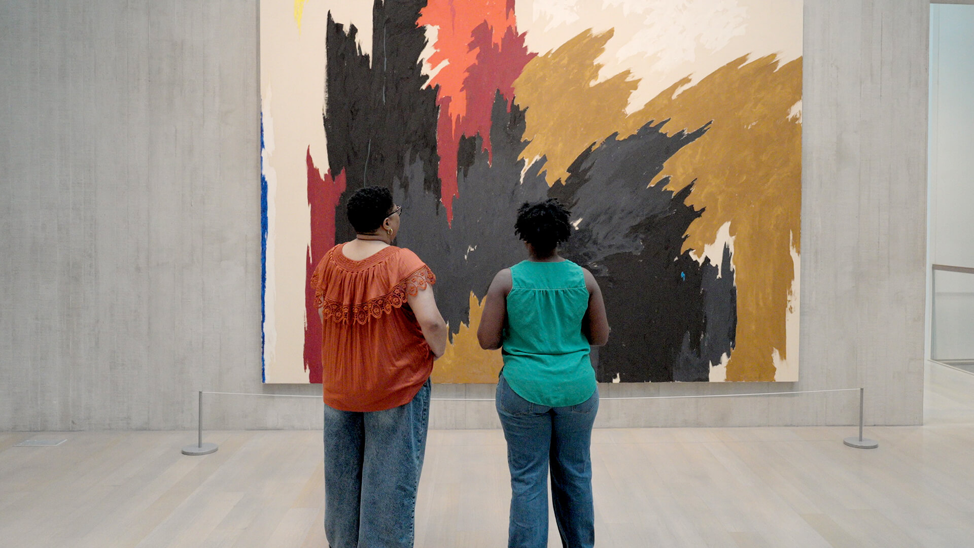 Two women look at a large colorful abstract painting on a concrete wall